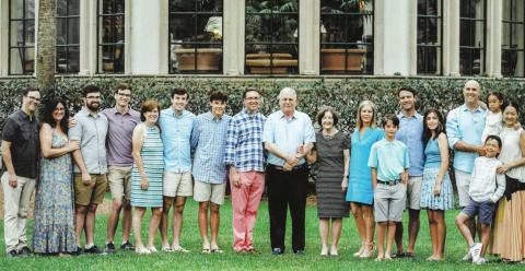 The extended Krugman family in casual summer attire standing in the grass in front of green shrubs and a building with large glass windows.