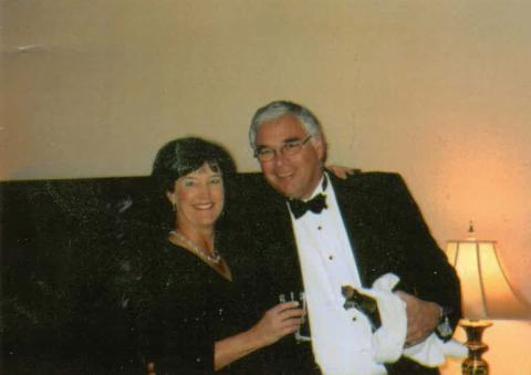 Smiling dark-haired woman in a black dress with pearls holds a glass, arm around a man in a tuxedo holding champagne wrapped in a white cloth.