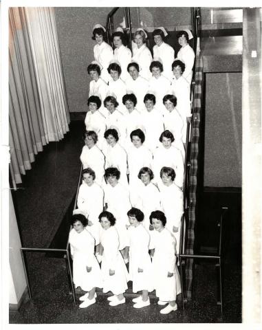 Black-and-white photo of 27 women on a staircase wearing 1960s-style white nurse uniforms and caps.