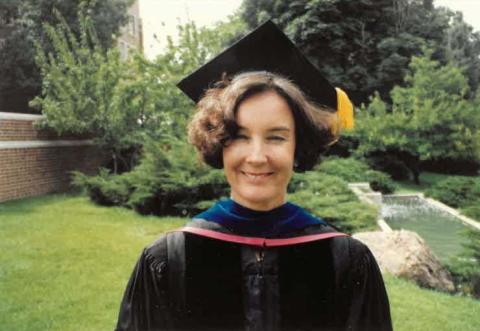 Smiling dark-haired woman in a black PhD cap and gown standing on grass with a brick wall and garden behind her.