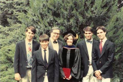 Smiling dark-haired woman in a black cap and gown holding a red diploma cover, surrounded by a man and four boys in suits.