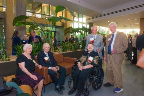 A woman and a man are seated on a couch, and another woman is seated in a wheelchair, while two men in blazers stand beside them in a plant-filled atrium lobby.