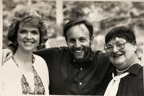A bearded man in a dark button-down shirt stands between two women, one wearing a floral dress and white cardigan, the other wearing round glasses, a polka-dot shirt, and a white scarf.