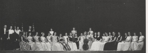 Seated orchestra of women in formal dresses arranged on a stage, with a few individuals standing at the edges.