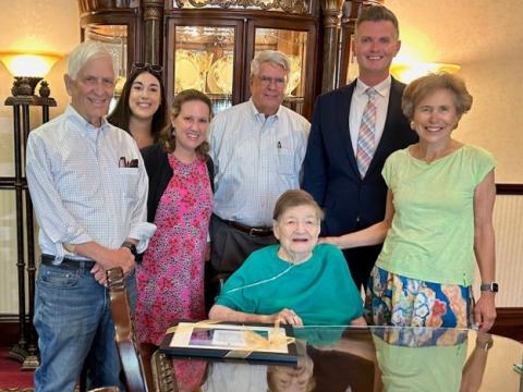 Three men and three women smiling and standing around Bonnie Camp, MD who is seated, wearing a teal shirt and an oxygen cannula. A framed award with a gold ribbon is sitting on a table in front of Dr. Camp.
