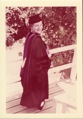 Bonnie Camp wearing a black graduation cap and gown standing on a wooden deck next to a railing with leafy trees in the background.