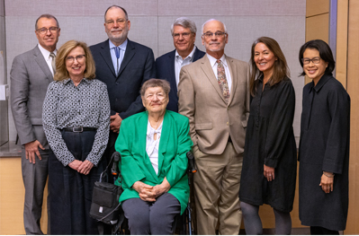 Five men and two women wearing business attire, smiling, and standing around Bonnie Camp, MD who is seated, wearing a green blazer.