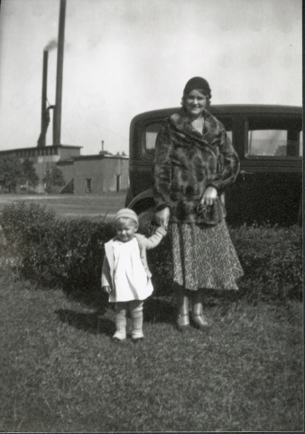 Black-and-white photo of a woman in a fur coat and patterned dress standing beside a young child in a light-colored coat. They hold hands in front of a 1940's car.