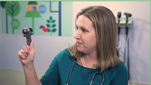 A female healthcare professional wearing a teal top and a stethoscope looks at a small elephant finger puppet on her raised finger in a pediatric clinic room.