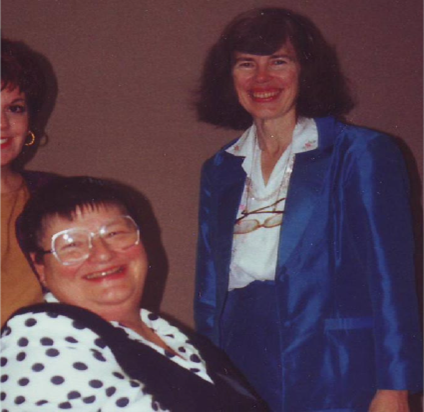 Three women smile at the camera. One woman wears round glasses and a black-and-white polka-dot shirt. Standing next to her, a woman in a royal blue pantsuit, while a third woman in a yellow shirt is partially visible on the left.