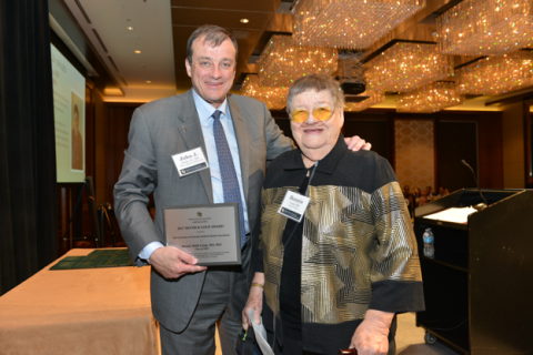 Dean John J. Reilly is in a gray suit and holds a framed plaque, while Dr. Bonnie Camp wears yellow-tinted glasses and a black-and-gold patterned jacket standing together in a ballroom.