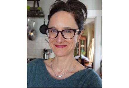 Mary Purdy smiling, wearing red lipstick, black glasses, a silver necklace, and green shirt. Her dark brown hair is pulled back into a bun.
