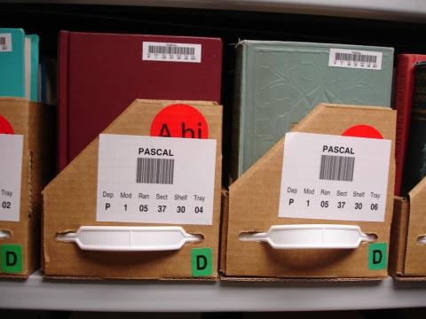Books stored in library shelving boxes with barcodes identifying contents.