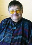 Portrait of a smiling Bonnie Camp, MD with short gray hair wearing round yellow-tinted glasses and a patterned blue-and-purple jacket with decorative buttons.