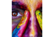Photograph close-up of a person's forehead, right eye, and nose, holding two fingers on either side of the bridge of the nose. The person's eye is brown and their skin has been painted with multiple bright colors, including red, pink, blue, purple, yellow, green, and orange.