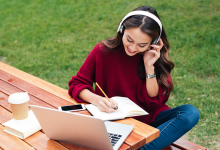 Young woman sitting at a table outside studying with headphones, a laptop, and a pen and notebook.
