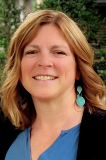 Lori Micho wearing a blue top and a black sweater, wearing turquoise earrings with greenery in the background.