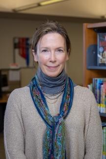 Wendy Kisicki wearing a light brown sweater with a blue turtleneck and colorful scarf with library bookshelves in the background.